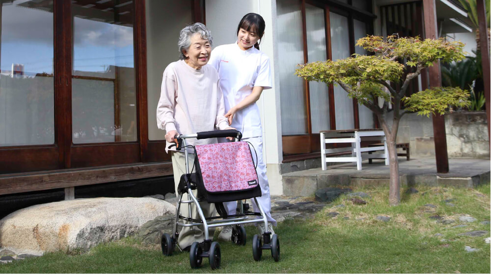 パーキンソン病のリハビリ効果で、屋外を歩く高齢女性と見守る施術者の様子