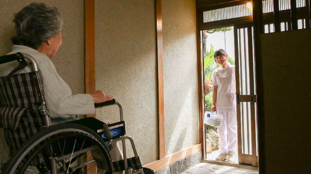 訪問鍼灸の施術者が自宅玄関に到着し、車椅子の利用者に挨拶する場面