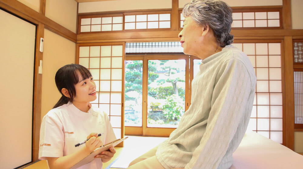 自宅の和室で訪問マッサージの施術者が利用者に問診し、体調や希望を丁寧に確認している様子。訪問マッサージの申し込みや相談の場面。