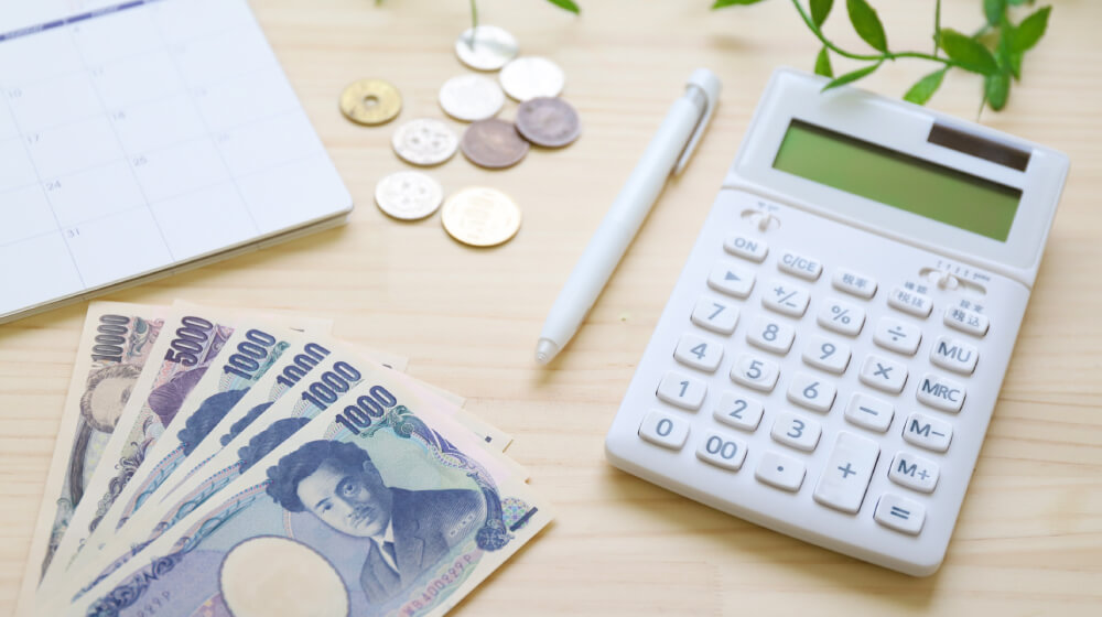 電卓と日本円の紙幣・硬貨、カレンダーが机に並ぶ。訪問マッサージの料金や自己負担額を計算・確認するイメージ。