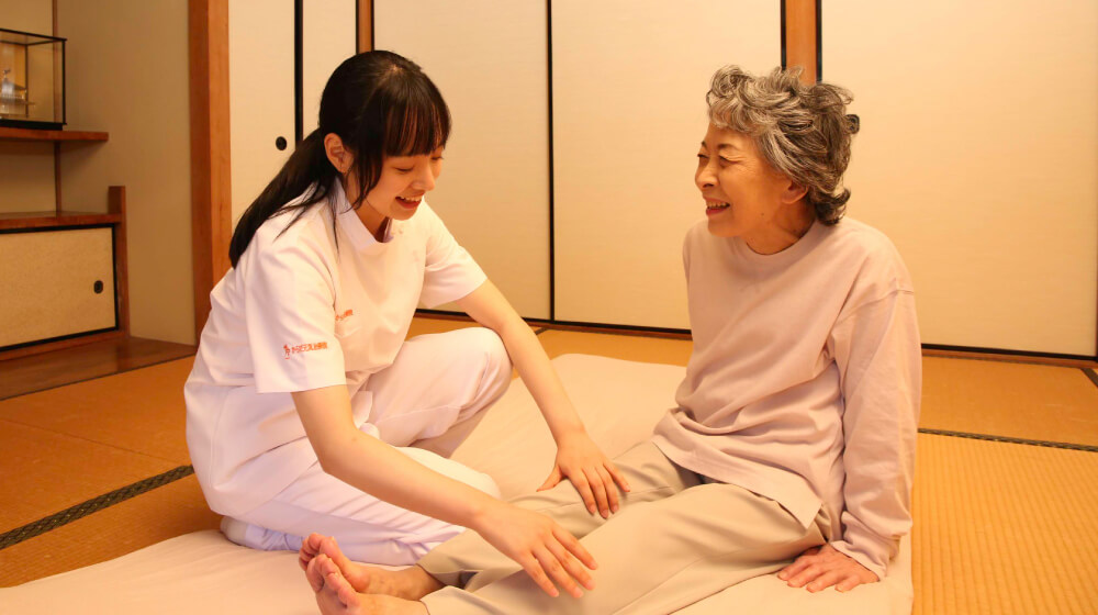 自宅の和室で施術者が利用者の脚を優しくマッサージしながら笑顔で会話している様子。訪問マッサージで心身をケアし安心して暮らすイメージ。