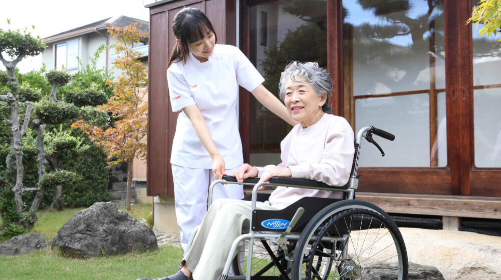 自宅の庭で車椅子に乗る高齢女性と、笑顔で寄り添う訪問サービスのスタッフ
